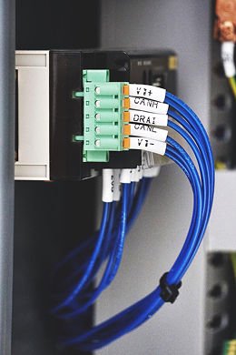 A close-up of blue networking cables connected to a terminal block, each labeled with tags such as V1+, CANH, DRAIN, and CANL inside an industrial control panel.