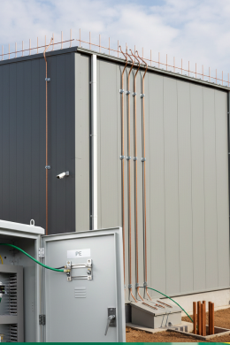 A grey industrial building with vertical copper pipes along its exterior wall. An electrical panel with open door and visible wiring is in the foreground, highlighting professional Electrical Services. A security camera is mounted on the building.