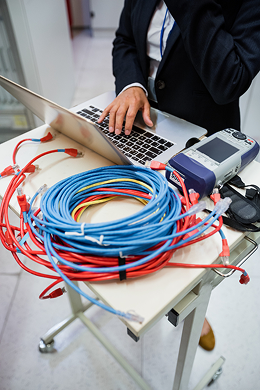 A person in business attire uses a laptop on a wheeled cart with coiled networking cables and electronic equipment in a server room or technical setting.