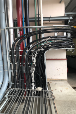 A bundle of black electrical cables and wires, expertly organized by electrical contractors, runs along a metal frame and bends around a corner in an industrial area with exposed pipes and concrete flooring.