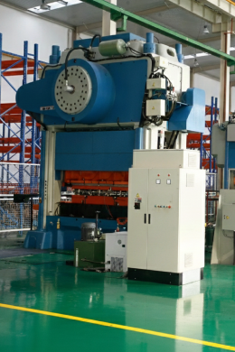 Large industrial press machine with blue and white components stands in a factory with green floors, metal shelves in the background, and control cabinets positioned nearby for integrated electrical services.