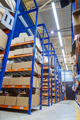Tall blue-metal shelves stacked with various cardboard boxes line a brightly lit warehouse aisle. The organized storage area highlights efficient Interior Services, with numbered signs and tiled floors illuminated by overhead lights.