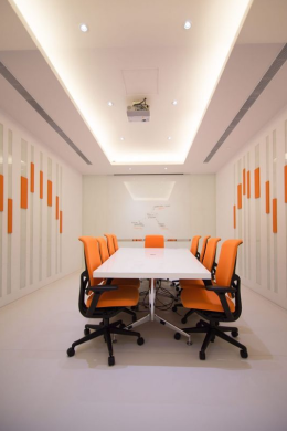 A modern conference room with a long white table surrounded by eight orange office chairs; white walls feature orange geometric patterns, and a projector is mounted on the ceiling, supported by reliable electrical services.
