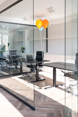 A modern office with glass walls showcases Interior Services expertise—empty black chairs at desks and three colorful balloons (yellow and orange) float near one desk as sunlight streams into the bright, minimalist space.