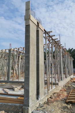 Concrete columns and wooden scaffolding rise at a building construction site, reflecting the dedication to civil service. The unfinished structure features exposed rebar, wooden supports, construction materials, and dirt beneath a partly cloudy sky.