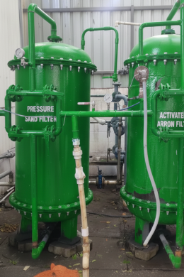 Two large green industrial filter tanks labeled Pressure Sand Filter and Activated Carbon Filter, essential for civil service water treatment operations, are connected by pipes and gauges, standing on a concrete floor in a plant setting.