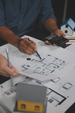 Two people review architectural blueprints at a desk, one holding a pencil and the other using a calculator—perhaps planning for a Civil Service building. Small model houses and additional floor plans are also visible on the table.